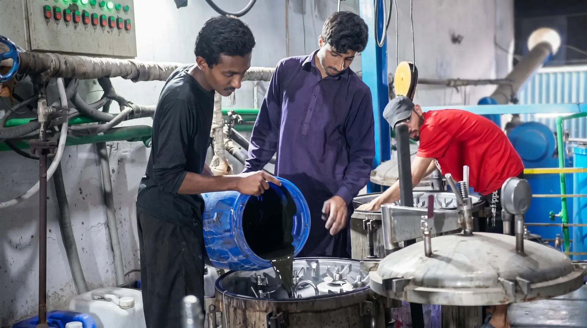 Three dye house operators at the kiers: one tipping dye liquor from a drum into an open vessel, one watching the pour, and one reaching across the lid of the kier beside it.